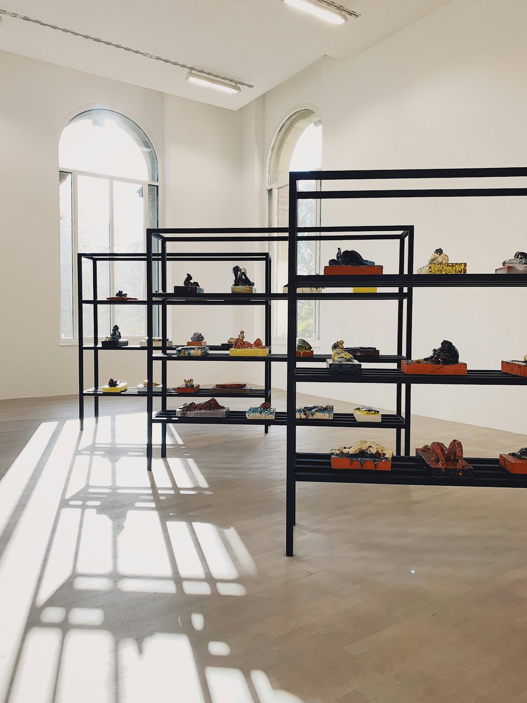 Colorful sculptural artworks arranged on black metal shelves in a gallery.